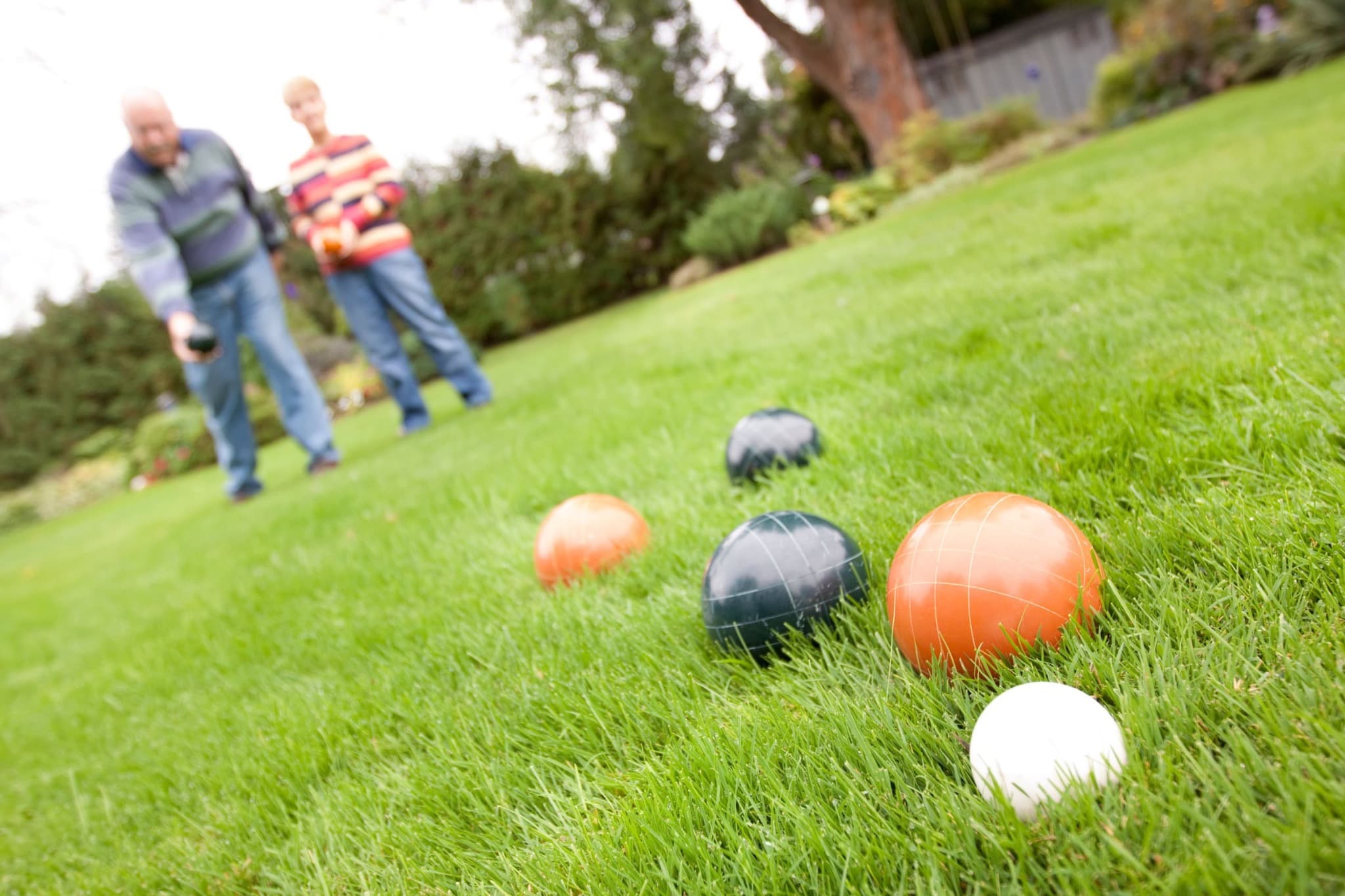 Bocce balls and a white pallino on a lawn, with two players in the background.