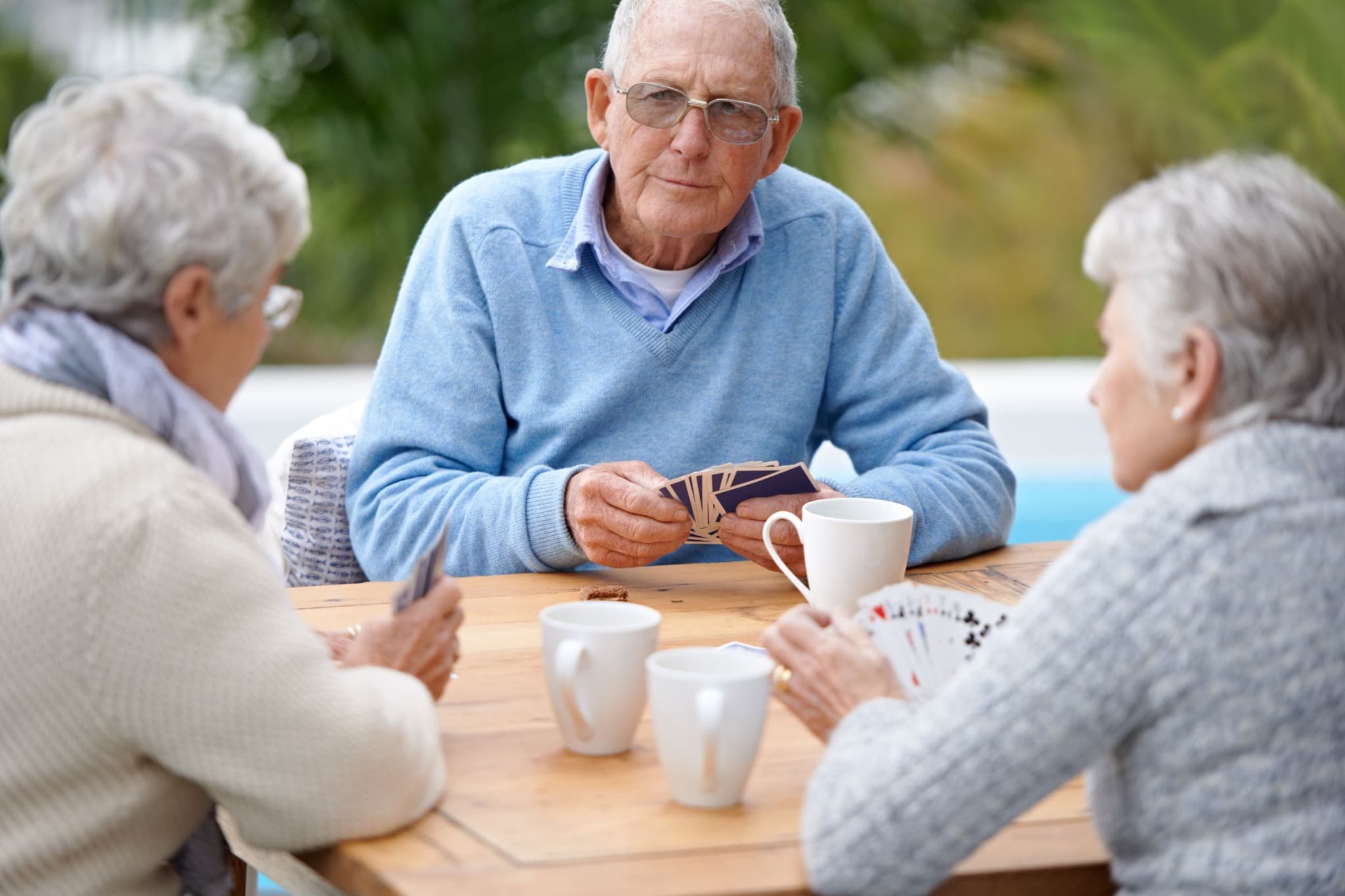 Three adults holding playing cards around an outdoor table.