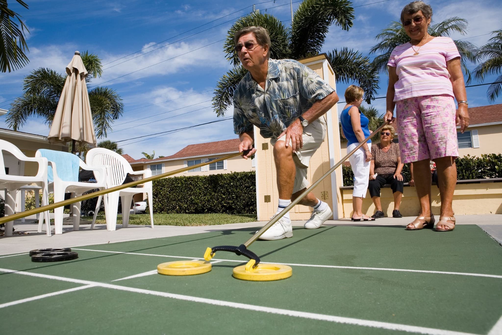 Two players using cues on an outdoor shuffleboard court with yellow and black discs.