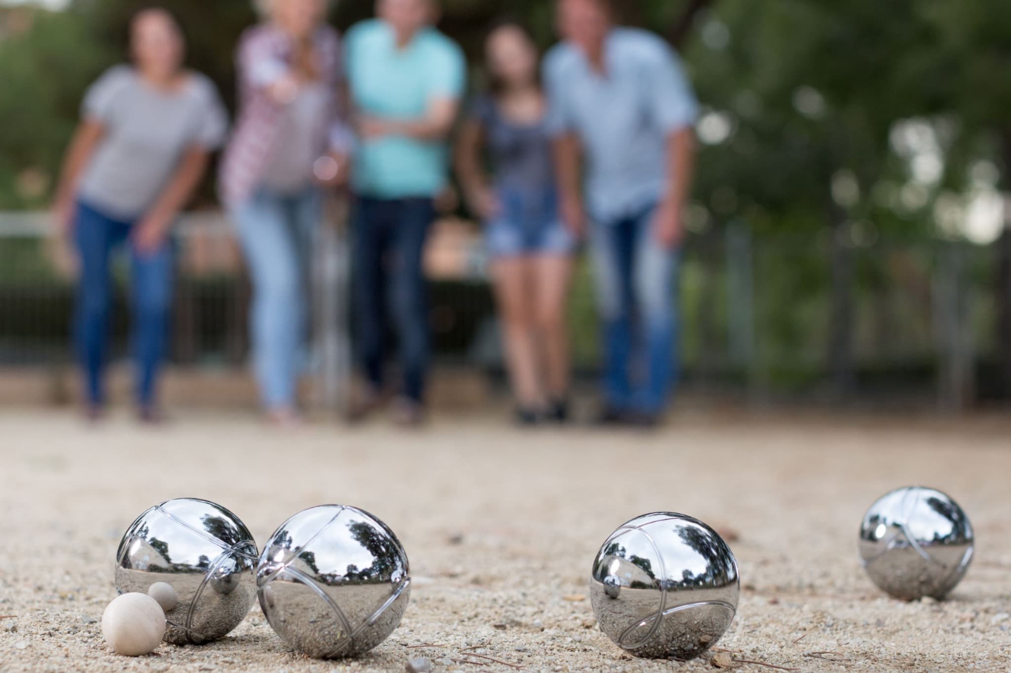 Steel pétanque boules and a small wooden jack on gravel, with players in the background.