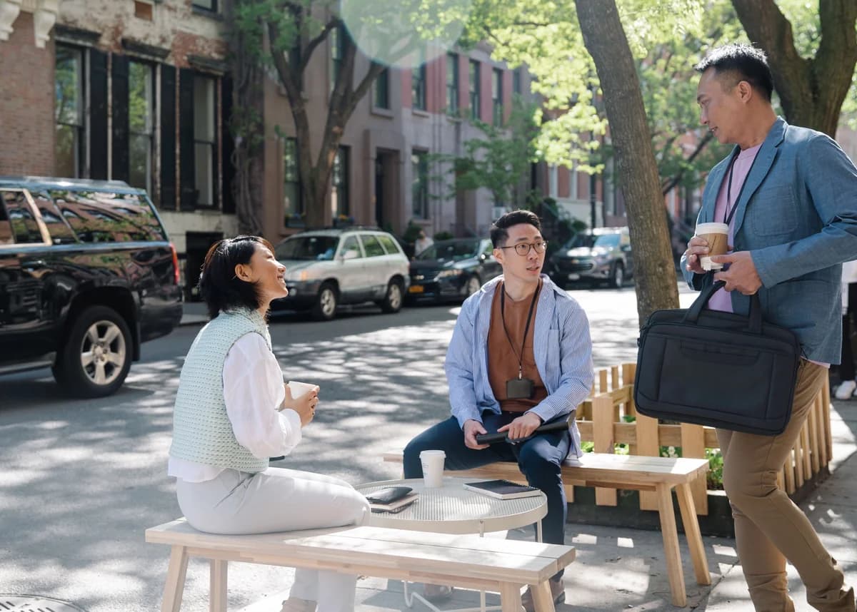 Three people chatting over coffee at an outdoor table on a tree-lined sidewalk