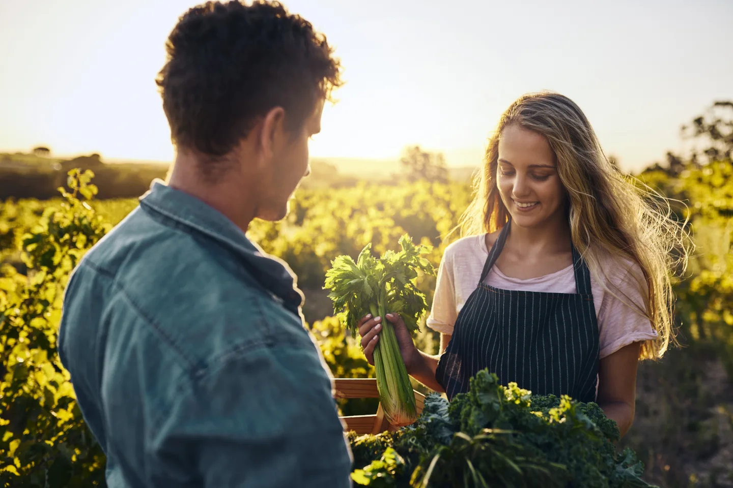 A woman in an apron showing fresh vegetables to a man outdoors.